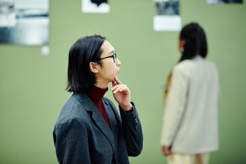 Young Asian man wearing eyeglasses standing in modern art gallery looking at photos at exhibition...
