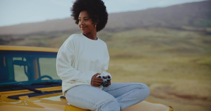 Beautiful young black african american woman relaxing and taking pictures at sunset with vintage camera on epic road trip, freedom and travel concept