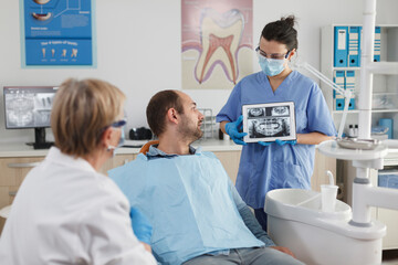 Obraz premium Stomatologist nurse with face mask holding tabet computer explaining teeth radiography to sick patient during dentistry consultation in stomatological office room. Team working at caries treatment