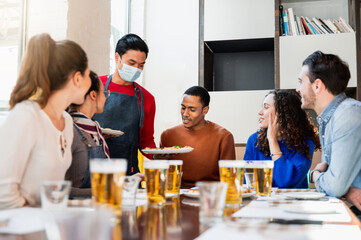 Asian young waiter wearing protective face mask serving pizza to a multiracial group of young friends sitting at table restaurant. People with happy smiling faces.