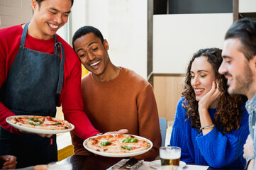 Multiracial group of friends sitting at table looking hungry the pizzas that Asian smiling male with apron is bringing to them