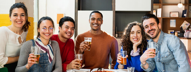 Horizontal banner or header portrait of Multiethnic group of young friends sitting at table eating pizza and drinking beer posing at camera with happy smiling expressions.