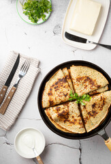 Flat lay thin cheese pancakes with greens, folded in a triangle in a cast iron pan, served with cutlery and butter on a concrete background