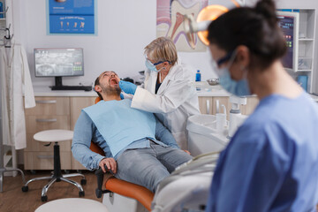 Fototapeta premium Specialist dentist senior woman using dental tools while checking patient mouth analyzing oral infection during stomatological consultation in dentistry office room. Concept of orthodontist team