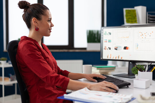 Successful businesswoman using desktop computer to analyze business data. Smiling entrepreneur looking at charts on pc screen in startup office. Employee in red shirt working on reports at desk.