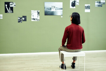 Rear view of unrecognizable young man sitting on stool in modern art gallery looking at black and white photos on green wall