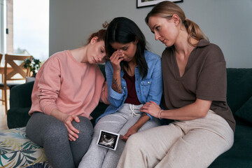 Mixed race woman holding ultrasound scan consoled by two caucasian friends