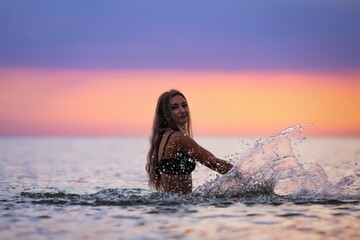 Fototapeta premium A girl with blond hair in a black swimsuit splashes to the sides while sitting in an estuary on a sunset background