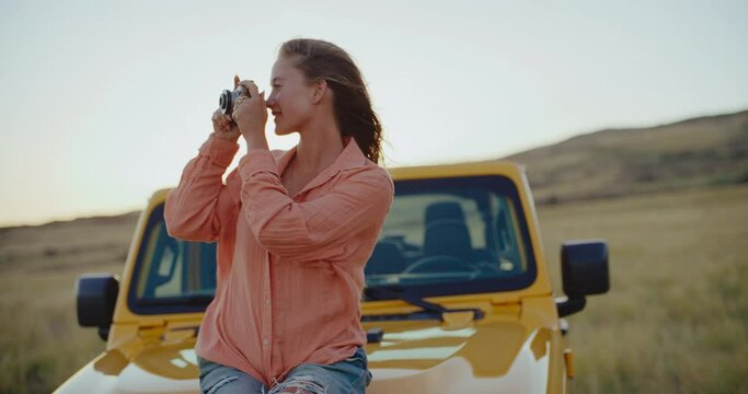 Beautiful young woman relaxing and taking pictures at sunset with vintage camera on epic road trip, freedom and travel lifestyle