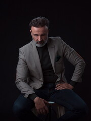 Portrait of adult businessman wearing trendy suit and sitting in modern studio on stylish chair against the black background