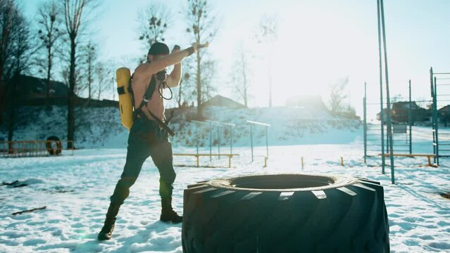 Muscular man with a naked torso with a drager on his back and in a full face mask hits a big tire with a sledgehammer. Slow motion