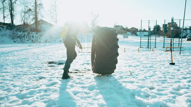 Bodybuilder with naked torso with drager on his back and wearing a full face mask does crossfit exercises and flips a big tire