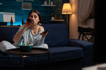 Young woman drinking beer from bottle and switching TV channels to find show or do movie selection. Casual adult enjoying alcoholic beverage and watching television program on couch.