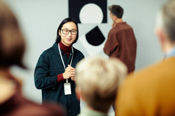 Young Asian man working in modern arts gallery standing in front of group of unrecognizable people speaking about exhibition - obrazy, fototapety, plakaty
