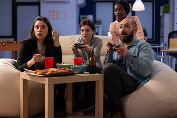 Office colleagues enjoying video games play with joystick on tv console at drinks party after hours. Cheerful friends having fun with technology, alcoholic beverage and snacks after work.
