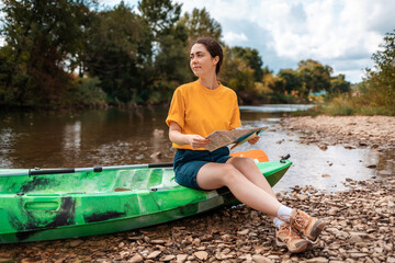 A happy young woman is sitting on a kayak, holding a paper map. Copy space. The concept of the World Tourism Day