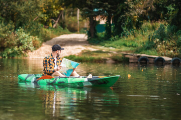 Fototapeta na wymiar Bearded caucasian man in shirt and cap sitting in kayak and holding a paper map. The concept of the World Tourism Day