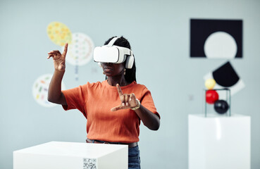 Young African American woman wearing VR headset looking at art objects at contemporary exhibition...