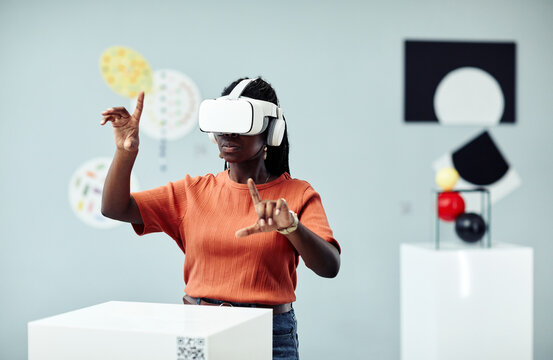 Young African American woman wearing VR headset looking at art objects at contemporary exhibition using augmented reality technology