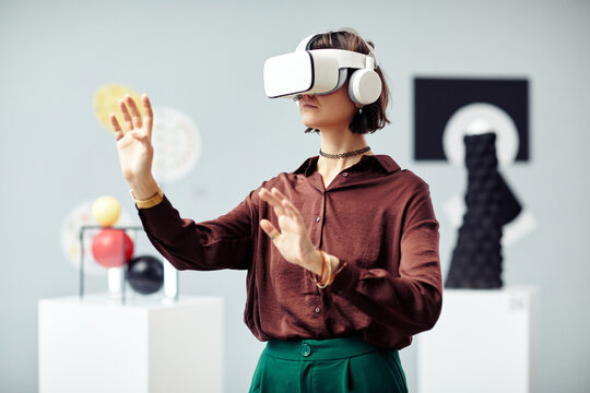 Young Caucasian woman wearing VR headset looking at art objects while visiting modern exhibition using augmented reality technology