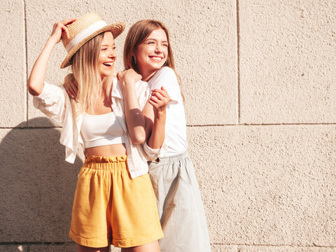 Two young beautiful smiling hipster female in trendy summer clothes.Sexy carefree women posing in the street near white wall in hat. Positive pure models having fun at sunset