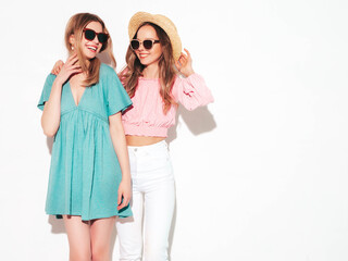 Two young beautiful smiling brunette hipster female in trendy summer dresses. Sexy carefree women posing near white wall. Positive models having fun. Cheerful and happy. In hats and sunglasses