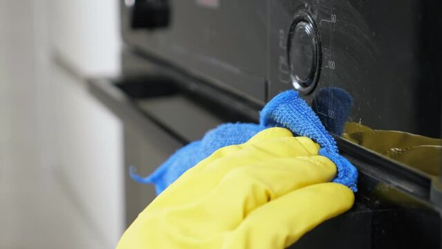 Man in yellow rubber gloves cleans oven wiping dust and stains with rag in kitchen. Housekeeper takes care of appliances condition at home closeup