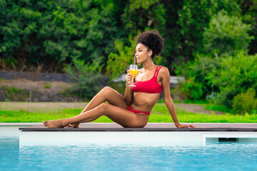 A young African American woman in a red bikini sunbathes by the pool while sipping a non-alcoholic...