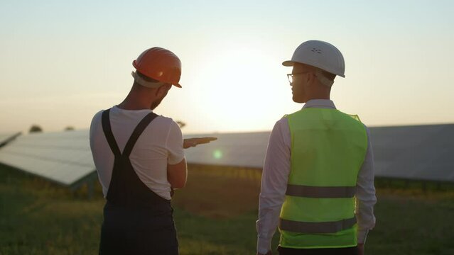 Back view of competent technician and qualified engineer discussing working project while standing together on field with solar panels. Amazing sunset on background.