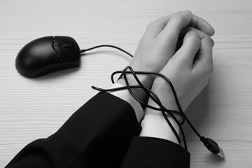 Closeup view of woman with her hands tangled in cable of computer mouse at wooden table, black and white effect. Internet addiction