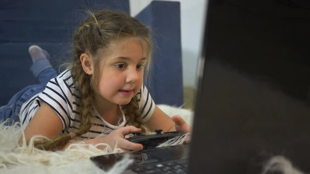 child in room, lying on couch, plays with computer console. gambling addiction of children. child online game on computer while at home. girl looks at monitor screen while operating remote control