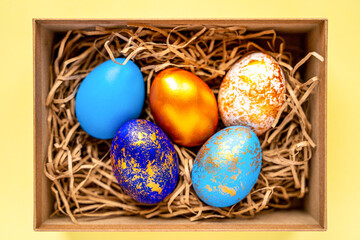 Fototapeta na wymiar colored golden, blue, blue easter eggs on a yellow background for easter and a place for text in a box and on a straw, happy easter