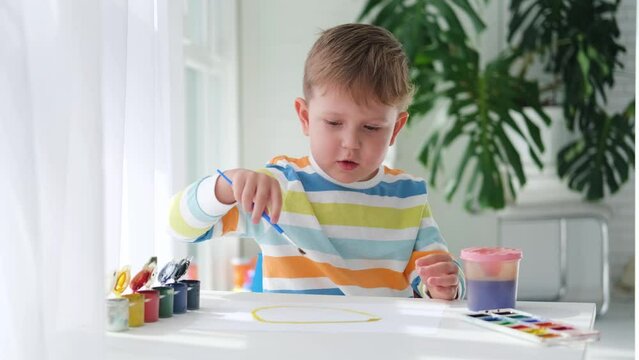 A little boy draws a picture with paints on a sheet of paper. The child is learning to draw. Home creativity.