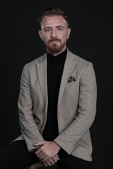 Portrait of adult businessman wearing trendy suit and sitting in modern studio on stylish chair against the black background