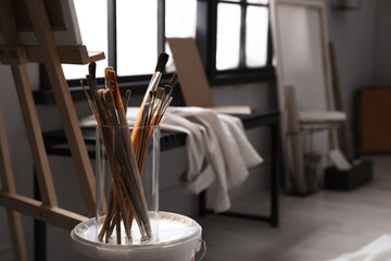 Holder with different brushes on bucket of paint near easel in artist's studio, space for text