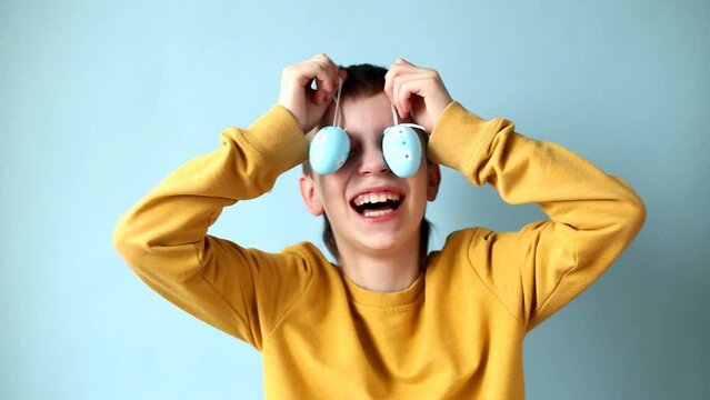 young smiling boy holding Easter eggs in his hand and covering his eyes with it on a blue background.