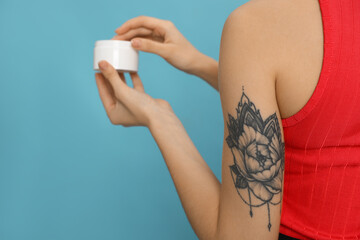 Woman with tattoo holding jar of cream against light blue background, closeup