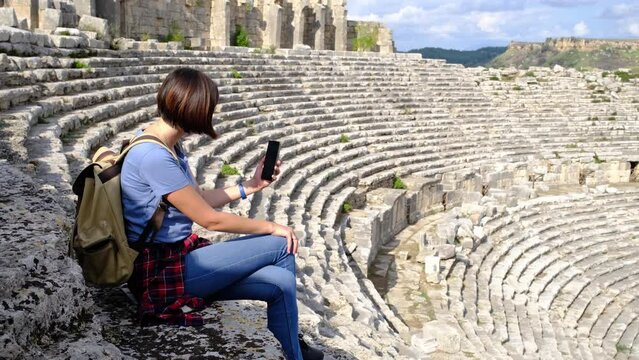 walk in Antalya Turkey on warm October afternoon. Pretty tourist woman with backpack at ruins of ancient city of Perge, large ancient amphitheater. taking photos of surrounding landscape on smartphone