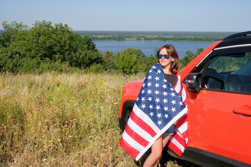 A young woman with an American flag stands near her car in nature.
