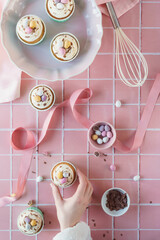 Flat lay Easter baking concept image. Easter theme cupcakes image with chocolate and baking equipment included in frame. Woman's hand reaches into frame to take a frosted cup cake. Pastel pink colours