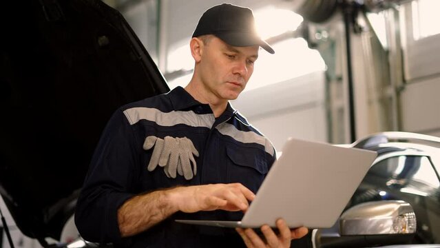 Car mechanic working on laptop in auto repair service, lean on a car, low angle view