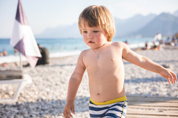 Cute child playing with yellow bucket and toy car on pebble beach. Happy toddler on sea coast. Summer family holiday vacation. Two years old little boy having fun. Summertime. Travel concept