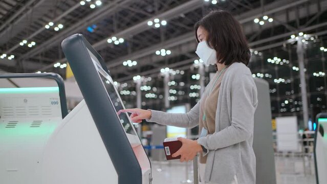 Woman wear mask using self service check-in kiosk at the international airport terminal. Touchscreen and see information on screen. Scan passport to record personal data. New normal travel
