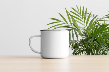 Enamel mug mockup on the wooden table with a palm plant.