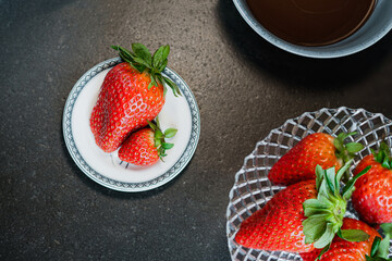 two strawberries on a white plate, in the background melted chocolate and more strawberries