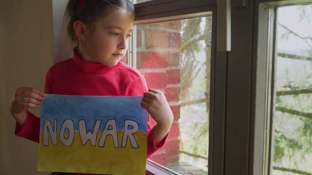 Refugee girl looking outside window and holding banner with inscription No war at blue yellow flag of Ukraine . Crisis, peace, stop aggression in whole world