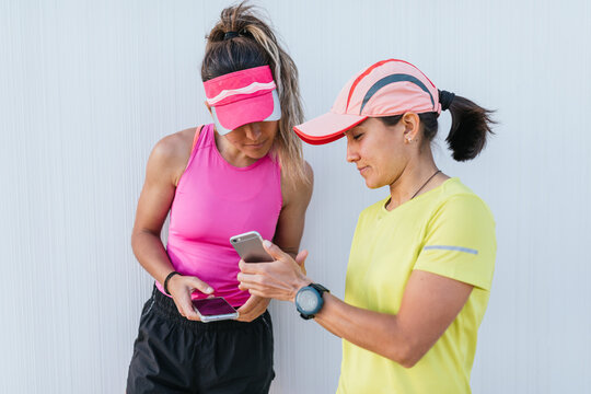 two beautiful athletic woman friends using app on mobile phone, looking at screen of smart phone on break between exercises