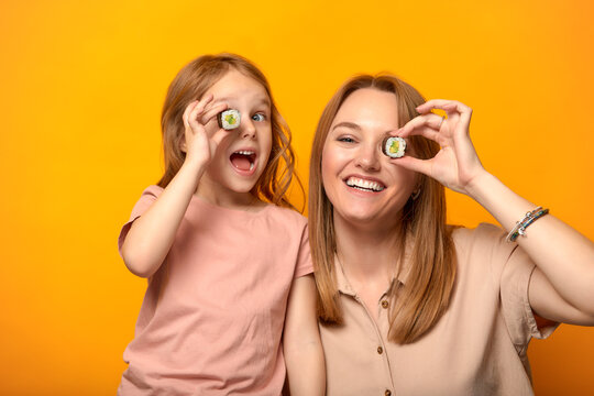 Funny mom with daughter holding sushi rolls in front of eyes on yellow background