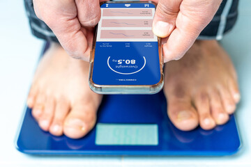 Mature man checking his body weight through an app on his smartphone while standing on the weight scale in the bathroom at home. High quality photo