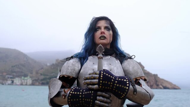 woman warrior in armor with sword and tears in eyes against backdrop of fortress on rock and sea. Warrior Soldier on Battlefield. War, Invasion, Crusade. Cinematic Historic Reenactment.